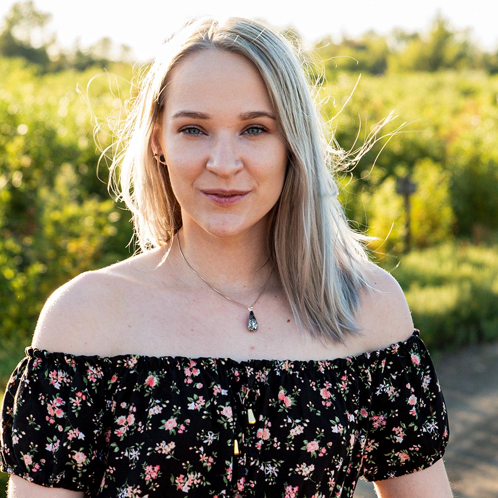 A woman wearing the petite obsidian teardrop memorial necklace poses in the sunlight in front of some green vegetation.