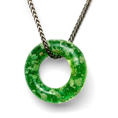 An image showing an Emerald Halo Ashes Pendant, a glass pendant infused with a small portion of ashes. The pendant is circular in shape, approximately 1.25" in diameter. It features vibrant colors and a delicate, handmade design. The pendant is suspended from a chain, which adds to its overall elegance. The image captures the personalized and heartfelt nature of the pendant, symbolizing the cherished memories and connection to a beloved person or pet.