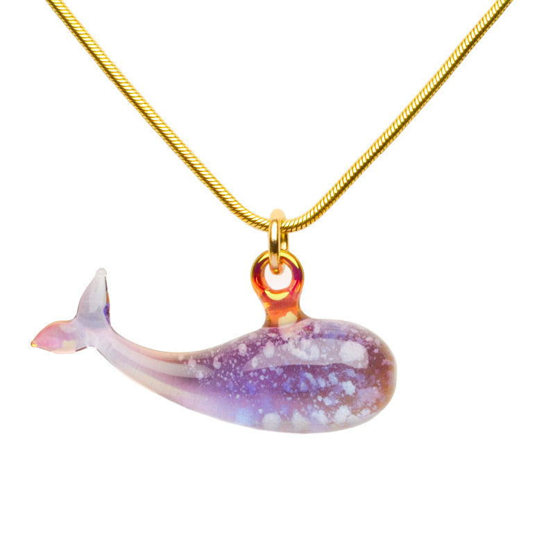 A purple/blue ash-infused cremation pendant on a gold chain features a graceful whale silhouette in glass, symbolizing strength and wisdom.